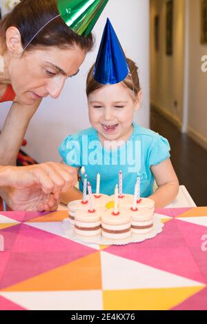 donna che illumina candele con accendino in mano sulla torta di compleanno su tovaglie colorate a casa, e tre anni bambino biondo guardando sorridente, con gr Foto Stock