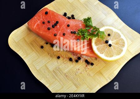 Una fetta di salmone crudo condito con prezzemolo, pepe, sale e limone su tritatutto in legno e fondo nero. Vista dall'alto Foto Stock