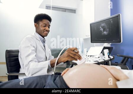 Primo piano di un medico africano uomo ecografo scansione di giovani donne incinte pancia con trasduttore a ultrasuoni che esegue ecografia ostetrica Foto Stock