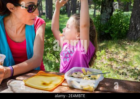 divertente bambina bionda di quattro anni che si diverte a giocare con i maccheroni, guardando la madre, mangiando la pasta da una scatola di plastica per pranzo in un tavolo da picnic in th Foto Stock