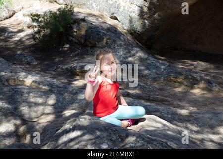 ritratto di quattro anni bionda carina ragazza, seduta sulla roccia all'aperto, guardando e facendo con le dita di mano il simbolo della vittoria Foto Stock