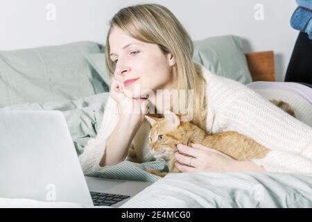 Donna casual che usa un computer portatile con il suo gatto rosso in camera da letto. Vita con animale domestico. Ombre Foto Stock