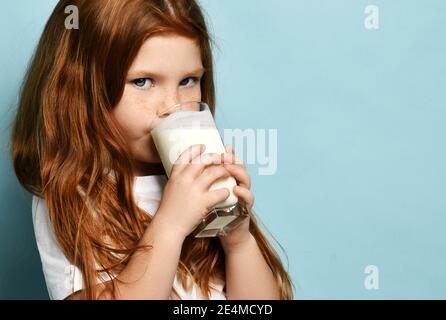 Piccola bambina dai capelli rossi che beve latte o yogurt kefir sorridendo gioiosamente su sfondo azzurro. Concetto di salute e dieta Foto Stock