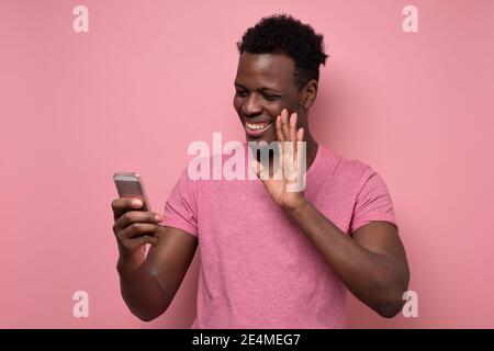 Gioioso sorridente giovane africano che tiene il cellulare in mano, agitando la mano durante la video chat. Scatto in studio Foto Stock
