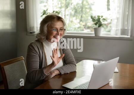 Donna matura sorridente che indossa occhiali che ondolano la mano, utilizzando un computer portatile Foto Stock