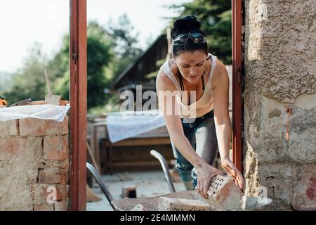 Costruttore femminile che carica una carriola con vecchi mattoni forati Foto Stock