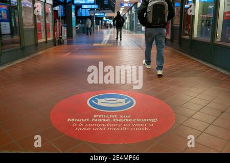 Fuoco selezionato al simbolo della maschera facciale bianca, e si prega di coprire il testo della bocca e del naso, sul segno rosso sul pavimento della stazione ferroviaria in Germania durante la socia Foto Stock