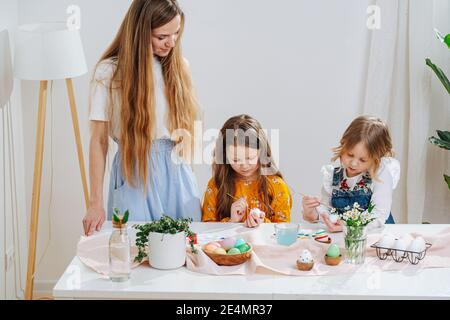 Due figlie dipingono le uova di pasqua dietro un tavolo, giovane madre che le guarda Foto Stock