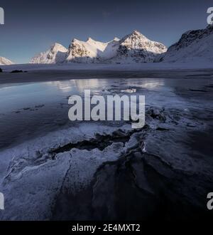 Tramonto invernale sulla spiaggia di Skagsanden, con interessanti texture nel ghiaccio speculare che riflettono le luminose montagne sullo sfondo. Foto Stock