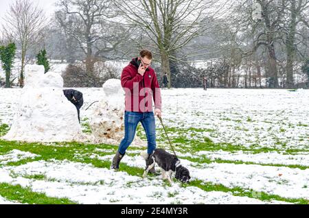 Un uomo cammina il suo cane mentre parla sul suo cellulare attraverso la neve in Prospect Park, Reading, Berkshire, UK. Foto Stock