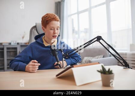 Ritratto di un adolescente dai capelli rossi che parla al microfono e utilizza un tablet digitale durante la lezione online o la lezione di e-learning, spazio di copia Foto Stock