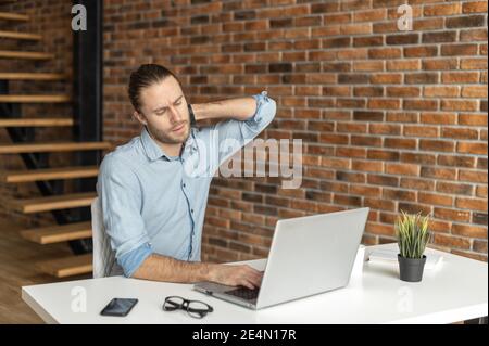 Frizzle ragazzo che usa il laptop per lavorare a distanza, un ragazzo stanco e scurchiato, che tiene indietro la testa e si sente un mal seduto alla scrivania in un ufficio moderno Foto Stock