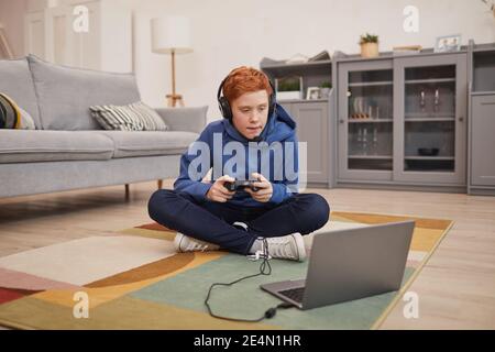 Ritratto a lunghezza intera di ragazzo adolescente con capelli rossi che gioca a videogiochi mentre si siede sul pavimento e tenendo il gamepad, spazio di copia Foto Stock
