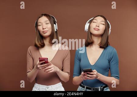 Giovani sorelle asiatiche in casualwear e cuffie godendo di relax musica e utilizzo degli smartphone mentre ci si trova davanti alla fotocamera Foto Stock