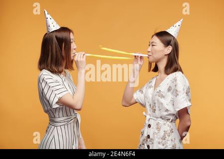 Felici giovani splendidi gemelli in abiti eleganti e berretti di compleanno in piedi di fronte l'uno all'altro e soffia fischi sfondo giallo Foto Stock