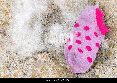 Un solo bambino perso calza sdraiato a terra. Il calzino è piccolo e viola, con una piccola quantità di neve accanto ad esso. Vista direttamente dall'alto. Foto Stock