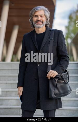 Uomo d'affari maturo con cuscinetti in elegante camice nero con rivestimento di fronte di macchina fotografica contro la scala del centro d'affari contemporaneo Foto Stock