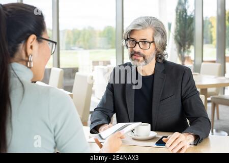 Uomo d'affari serio maturo in formalwear rispondendo alla domanda del suo giovane collega femminile alla riunione con una tazza di caffè al ristorante Foto Stock