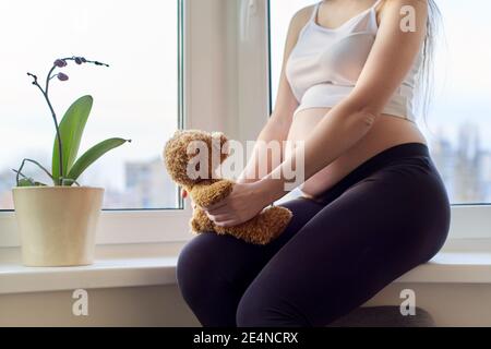 Donna giovane incinta felice con orsacchiotto giocattolo, a casa vicino alla finestra Foto Stock