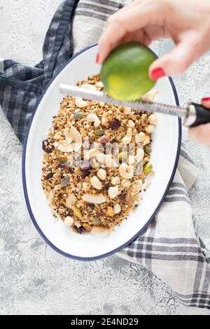 Le mani della donna grattugiano una calce su un vassoio bianco metallico pieno di deliziosa granola su un tovagliolo rustico su sfondo bianco e grigio. Foto Stock