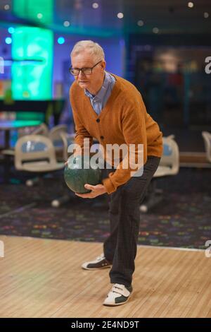 Ritratto verticale a tutta lunghezza di un uomo anziano attivo che gioca a bowling, in piedi su una corsia pronta a lanciare mentre ti godi l'intrattenimento Foto Stock