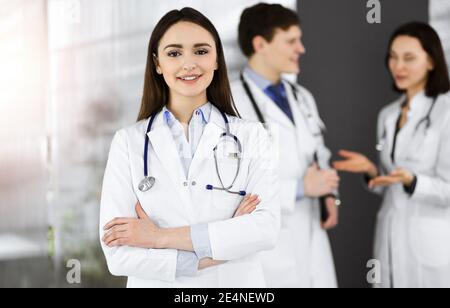 Sorridente donna-medico intelligente è in piedi con le braccia incrociate in clinica soleggiata insieme ai suoi colleghi sullo sfondo. Servizio medico perfetto Foto Stock