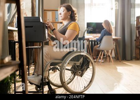 Giovane donna d'affari con disabilità in sedia a rotelle seduta dalla macchina xerox e premendo il pulsante di avvio durante l'esecuzione di copie di documenti Foto Stock