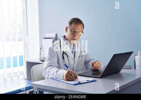 Medico anziano che usa il laptop in ospedale che guarda il webinar medico, che scrive il rapporto. Foto Stock