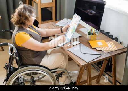 Giovane economista femminile in casualwear seduto in sedia a rotelle di fronte di computer monitor e prendere appunti durante l'analisi dei dati finanziari Foto Stock