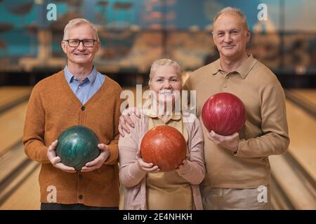 Vita in su ritratto di tre persone sorridenti senior che tengono le palle da bowling e guardando la fotocamera mentre si gode l'intrattenimento attivo al bowling, cop Foto Stock