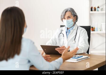 Medico femminile senior di mezza età in un camice da laboratorio e una maschera facciale seduta alla scrivania, tenendo un touchpad, discutendo i risultati medici con paziente, giovane Foto Stock