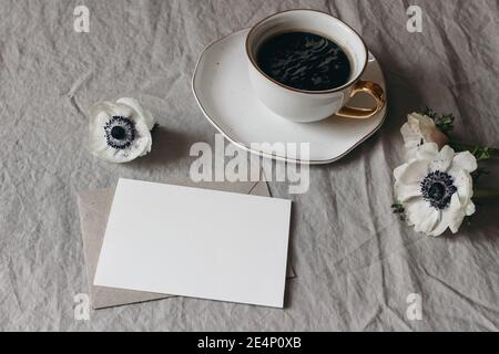 Scena mock-up di cancelleria per matrimoni primaverili. Biglietto d'auguri vuoto, busta su sfondo tovaglia di lino beige. Fiori bianchi di anemone e tazza di caffè Foto Stock