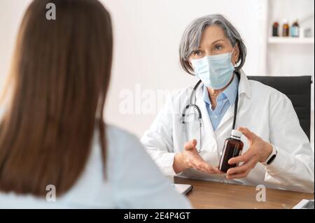 Senior maturo medico femminile in una uniforme medica e maschera protettiva viso seduta alla scrivania e parlare con il suo paziente, spiegando la diagnosi e. Foto Stock