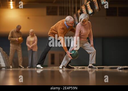 Vista laterale grandangolare presso la moderna coppia senior che gioca a bowling insieme mentre ti godi l'intrattenimento attivo nella pista da bowling e nello spazio fotocopie Foto Stock