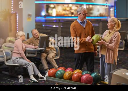 Ritratto di una coppia sorridente senior che gioca a bowling con gli amici in background mentre ti godi l'intrattenimento attivo al bowling, copy space Foto Stock