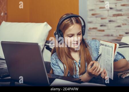 Felice giovane adolescente ragazza in cuffia che parla guardando il portatile e mostrando libro, ragazza studente che parla con videochiamata, insegnante insegnante tutorin Foto Stock