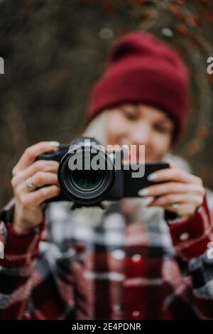 Ritratto di giovane donna che usa la macchina fotografica nella foresta. Ragazza che riprende e sorride, signora in camicia trendy e berretto rosso. Foto Stock