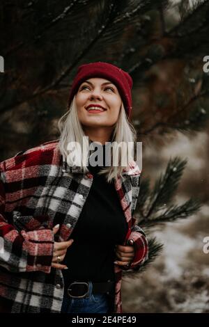 Ritratto di giovane bella donna in foresta, stagione invernale. Bella ragazza tipo sorridente, signora in camicia trendy e berretto rosso. Foto Stock
