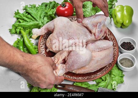 Mano dell'uomo che tiene il pollo intero crudo su piastra di ceramica con verdure fresche e spezie Foto Stock