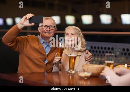 Ritratto di coppia senior moderna scattando foto selfie mentre beve birra al bar e godendo la notte con gli amici, spazio copia Foto Stock