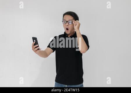 WOW faccia del tuo uomo asiatico ha scioccato ciò che vede nello smartphone su uno sfondo grigio isolato. Indonesia uomo indossare camicia nera isolato sfondo grigio. Foto Stock