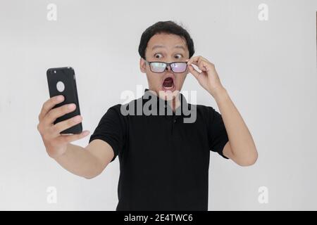 WOW faccia del tuo uomo asiatico ha scioccato ciò che vede nello smartphone su uno sfondo grigio isolato. Indonesia uomo indossare camicia nera isolato sfondo grigio. Foto Stock