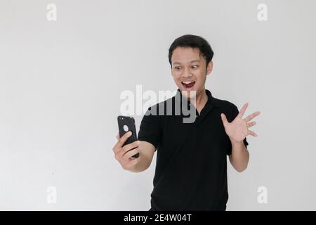 WOW faccia del tuo uomo asiatico ha scioccato ciò che vede nello smartphone su uno sfondo grigio isolato. Indonesia uomo indossare camicia nera isolato sfondo grigio. Foto Stock