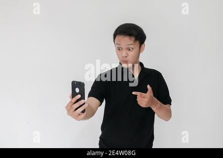 WOW faccia del tuo uomo asiatico ha scioccato ciò che vede nello smartphone su uno sfondo grigio isolato. Indonesia uomo indossare camicia nera isolato sfondo grigio. Foto Stock