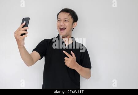 WOW faccia del tuo uomo asiatico ha scioccato ciò che vede nello smartphone su uno sfondo grigio isolato. Indonesia uomo indossare camicia nera isolato sfondo grigio. Foto Stock