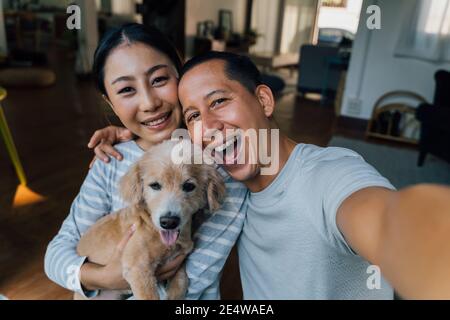 Giovane coppia asiatica adulta che tiene un cucciolo prendendo un selfie da un telefono con casa interna in background. uomo e donna maturi di 30 anni con cane animale domestico che scatta una foto della famiglia. - felice ritratto di gruppo Foto Stock