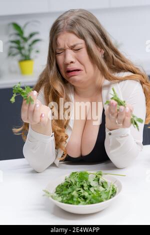 Fat ragazza non felice tenendo fresco insalata casting. Giovane donna dal corpo curvo con lunghi capelli biondi seduta sulla cucina moderna. Concetto di dieta e nutrizione Foto Stock