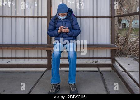 Un uomo maturo in maschera medica si siede in autunno ad una fermata di trasporto pubblico e attende un autobus, tram, filobus. Utilizzando lo smartphone, cerca informazioni Foto Stock