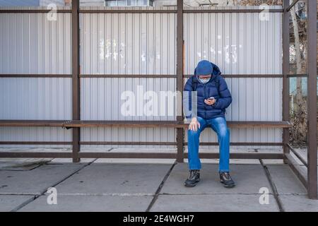 Un uomo maturo in maschera medica si siede in autunno ad una fermata di trasporto pubblico e attende un autobus, tram, filobus. Utilizzando lo smartphone, cerca informazioni Foto Stock