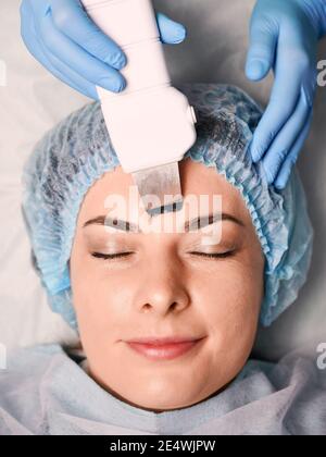 Primo piano di mani estetiste in guanti sterili utilizzando scrubber ultrasonico durante la pulizia della faccia del cliente femminile. Donna che ha pulizia profonda procedura di cura della pelle in salone di bellezza. Concetto di trattamento della pelle Foto Stock